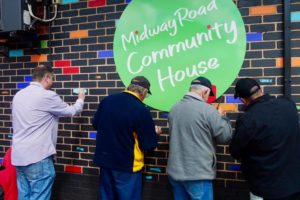 A grey brick wall with the community centre's logo. A small group of people are painting the bricks on the wall in different colours.