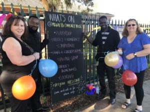 People stand around a blackboard holding balloons and smiling at the camera. The blackboard reads 'what's on @ Midway community house'