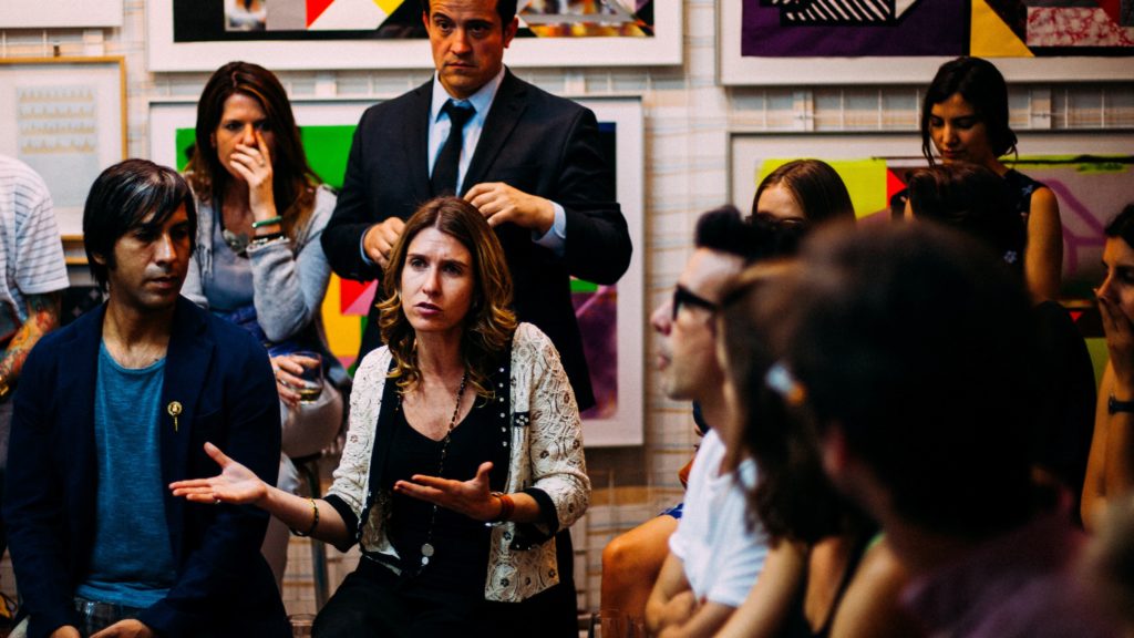 A group of people are listening to one person speaking who is facing the camera. The person's arms are held out as she explains to the group