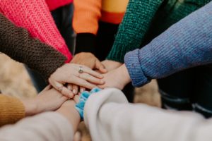 A cluster of hands coming together in a huddle, they are all wearing different coloured jumpers