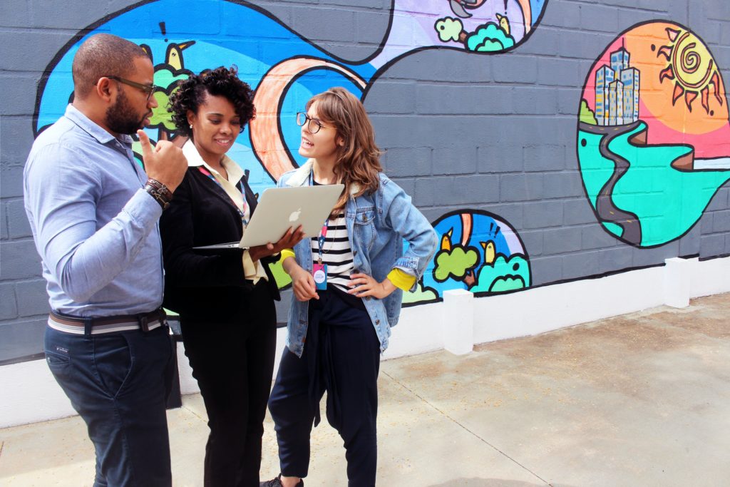 three people standing in front of a colourful mural. The person in the middle is smiling and holding a laptop while the two other people speak to each other